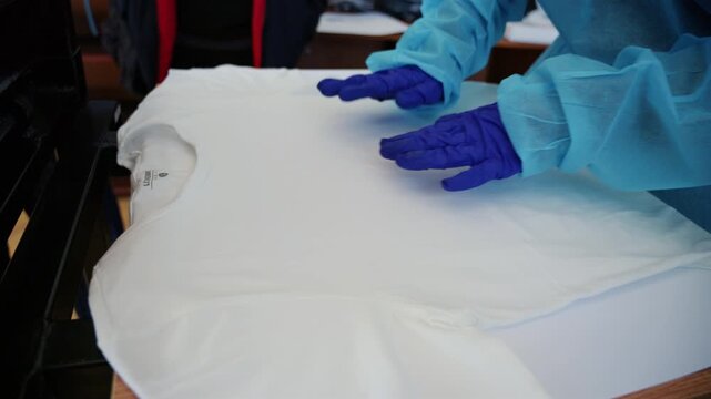 Close up footage of workers wearing protective gloves carefully positioning a white T-shirt on a screen printing press before printing a UTM logo