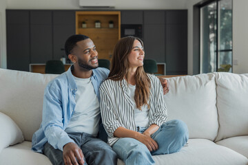 Obraz premium Happy young multiracial couple sitting on a cozy sofa in a modern living room, smiling and relaxed as they lean together, enjoying domestic comfort and quiet connection