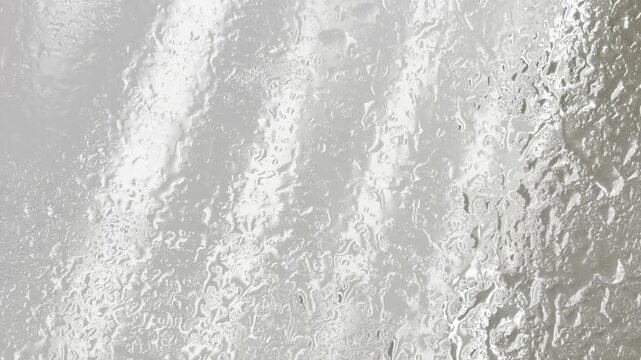 Water droplets condensing and collecting on a foggy window glass surface, creating a beautiful abstract background with calming rain like patterns and gentle light reflections