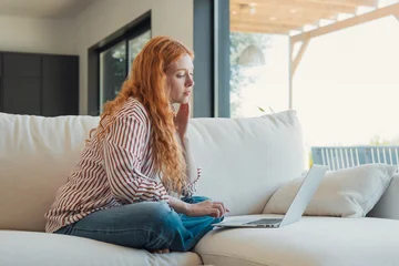 Fototapeten Lotusblume Redhead woman sitting in a lotus position on a comfortable couch, concentrating on her work using a modern laptop in a bright contemporary living room  © Daniel