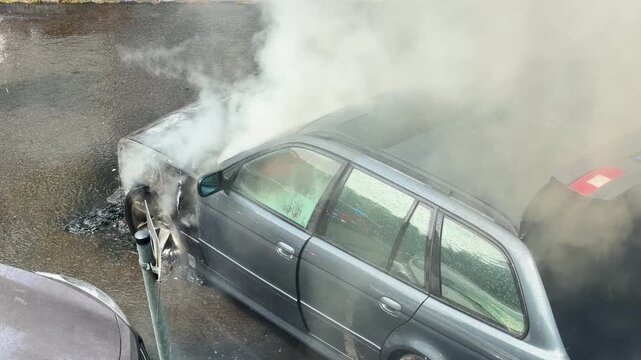A water stream hits a smoke covered station wagon after an engine fire. White steam, wet asphalt and charred panels show an active urban emergency response.