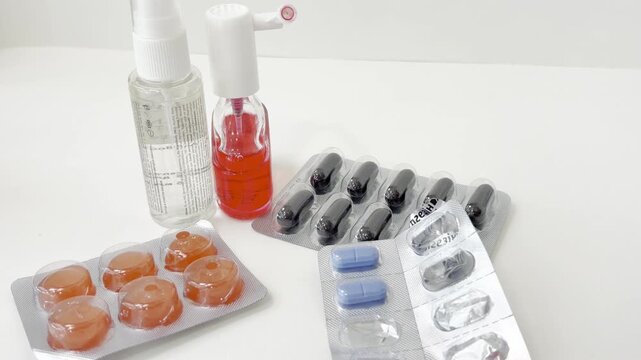 Person taking pills from blister pack for cold and flu treatment isolated on white background with throat spray, cough drops, and various medications for illness relief