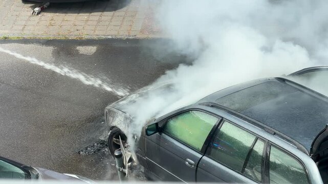 Thick white smoke obscures a burned station wagon as water enters the engine area. The wet roof, open hatch and haze emphasize the intensity of suppression work.
