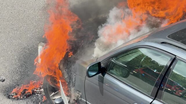 Orange flames burn around the front of a parked station wagon on a city street. Smoke, soot and heat show an intense vehicle fire before suppression.