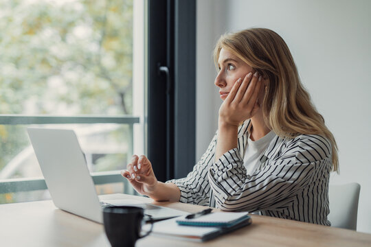 Woman feeling unmotivated and procrastinating, sitting at a desk with an open laptop, looking out a window and distracted from her work, showing burnout and mental fatigue