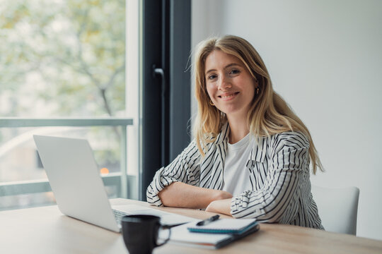 Young businesswoman smiling at laptop on bright home office desk, focused on remote work and online business tasks, conveying confidence, flexibility and productivity