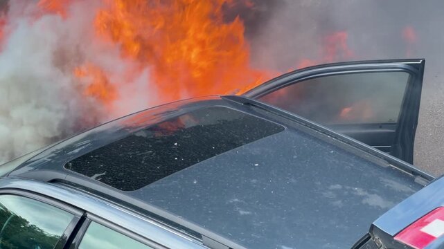 Flames and smoke spread across the roof and open door of a burning vehicle. The close view captures heat, soot and the intensity of the car fire.