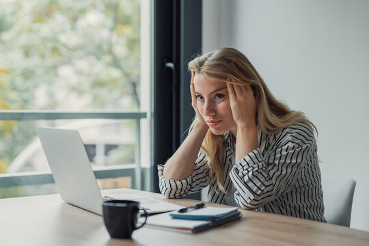 Blonde woman sitting at a desk with a laptop and coffee, holding her head in frustration and looking away from the screen, feeling overwhelmed and stressed during her remote work day