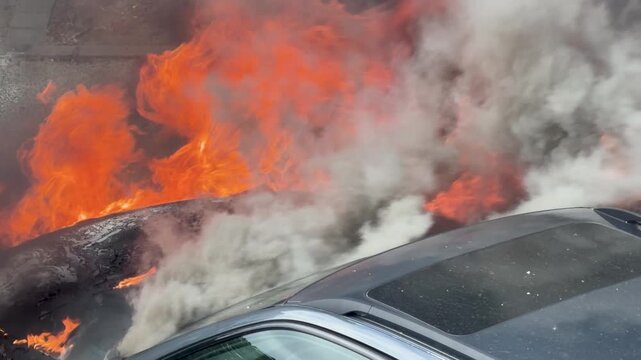 Close footage shows flames and white smoke pouring from a burning engine bay. The vehicle roof and charred hood frame the severe street fire.