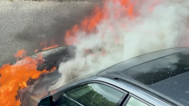 Massive orange flames consume the side and front of a parked car. Dark smoke, heat shimmer and close framing emphasize a severe urban vehicle fire.