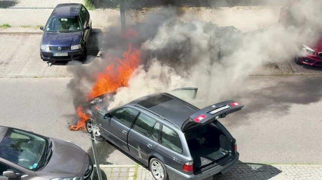Heavy black and white smoke rises from a burning station wagon on a city street. Flames at the front wheel and open hatch show an active vehicle fire.