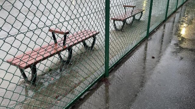  雨の日に金網フェンスの向こう側に佇む誰もいない公園のベンチ（ノスタルジー・背景素材）