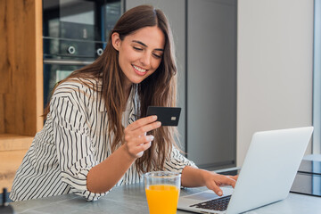 Fototapeta premium Young woman in a kitchen smiling at her laptop while holding a credit card, shopping online and managing digital banking from home with comfort and convenience