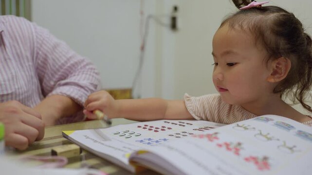 A focused toddler receives one-on-one tutoring from her mother, carefully practicing counting and writing in a workbook for her early development.