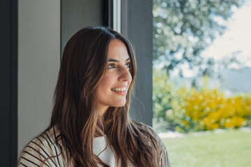 Woman with long brunette hair smiling while looking away through a window, experiencing emotions of...