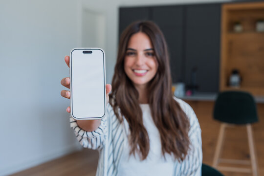 Woman smiling and holding a modern smartphone with a blank white screen as a mock-up, presenting an app or service in a casual home setting with blurred background space for copy