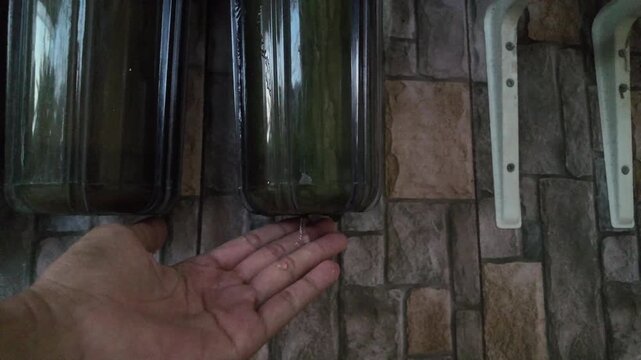 High angle view of a human hand placed underneath a dirty 10-inch water filtration housing cylinder to catch leaking water droplets in real time, showing damaged filtration system checkup.
