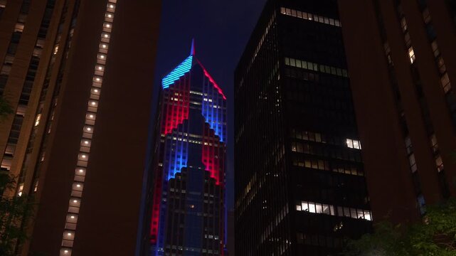 Tall Chicago skyscraper glowing with red and blue lights between dark high-rise buildings at night. Dramatic urban cityscape with illuminated windows and modern architecture.