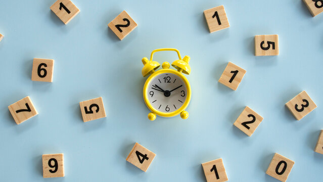 Flat lay of a small yellow alarm clock surrounded by scattered wooden number tiles on a plain background, concept of time management, deadline, and countdown.