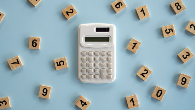 Top view of a white electronic calculator centered among scattered wooden number tiles on a light blue background, concept of accounting, mathematics, and budget planning.