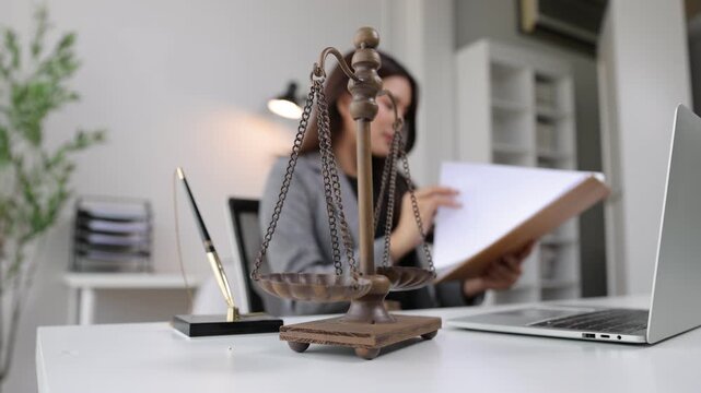 Scales of justice on a desk with a female lawyer working in the background. Close-up view of the scales of justice on a desk. A serious female lawyer works in the blurred background of her office.