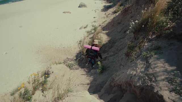 Young adult woman is hiking down a sandy path surrounded by grass towards the beach while carrying equipment on a sunny day.