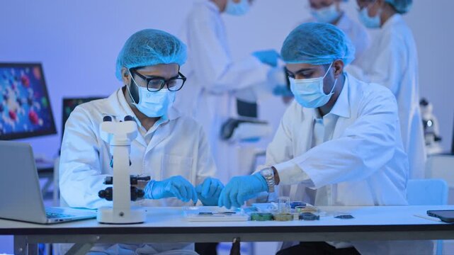 Male researchers in hairnets and masks examine slides at a microscope under atmospheric blue lighting, biomedical, biotech and laboratory concept.