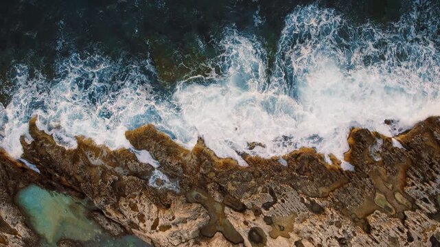 Aerial top view of swirling sea foam outlines layered rocks along tidal boundary