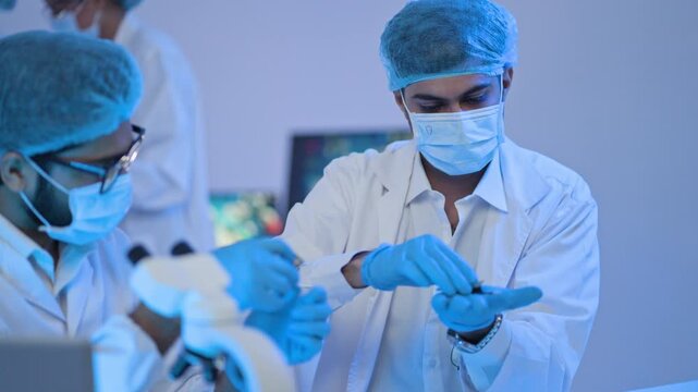 Male researchers in hairnets and masks inspect a tiny specimen with precision instruments under blue light, perfect for microelectronics and biotech themes.
