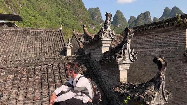 Slow motion shot of a male traveler with a backpack sitting on a traditional undulating tiled roof of a Ming‑and‑Qing‑era house, overlooking a scenic karst mountain landscape in Xingping Fishing Villa
