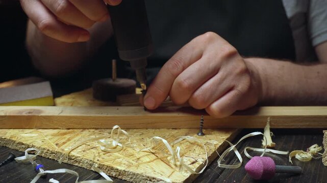 Skilled craftsman uses electric screwdriver to fasten wooden pieces together on a workbench surrounded by shavings and tools in a workshop setting