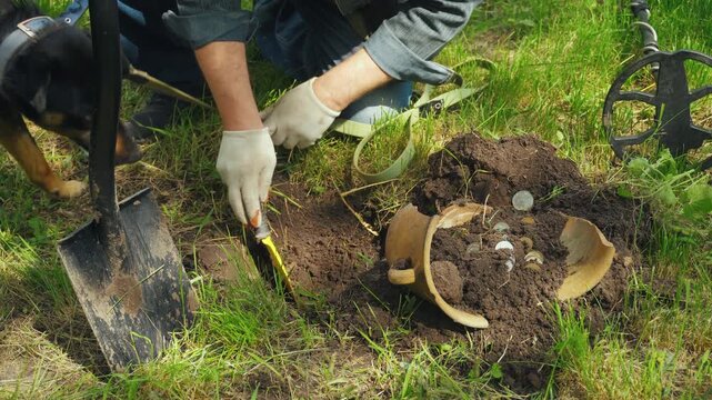 Man with dog digs in the ground using a trowel, uncovering coins and pottery fragments in a grassy area with a metal detector nearby