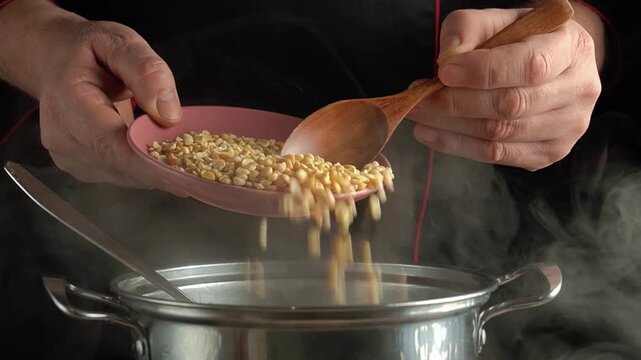 Chef pours lentils from a pink plate into a steaming pot using a wooden spoon, showcasing the cooking process in a kitchen setting