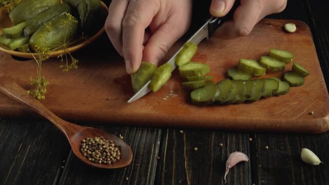 Person in a kitchen slices pickle on a wooden cutting board with a knife, while garlic cloves and a bowl of pickles are visible in the rustic kitchen environment