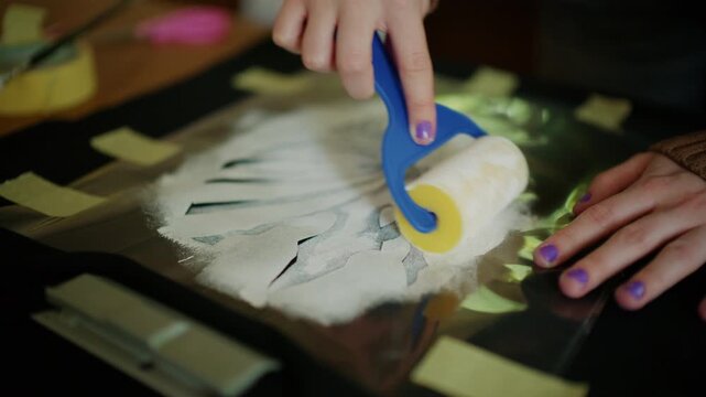 Hands using a craft roller to evenly spread adhesive material over a stencil design
