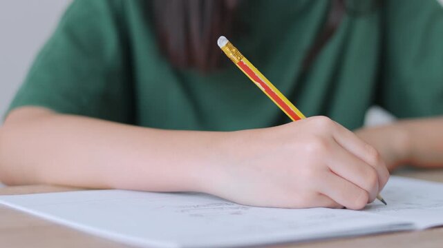 Close up selective focus of a student's hand holding a pencil, writing in a notebook. Bright daylight for the workspace, representing academic concentration,homework, and childhood education concepts.
