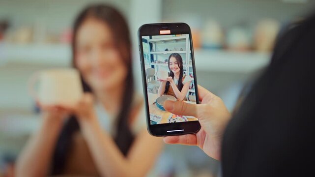 Close up of hand holding smartphone to record video of happy young Asian woman showing ceramic mug, creative female artisan vlogging in pottery studio, content creator, small business marketing