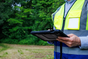 Obraz premium Close-up hands of ecologist or forestry engineer holding digital tablet in protective rugged case during field research in green forest. Data logging, environmental monitoring and tree mapping outdoor