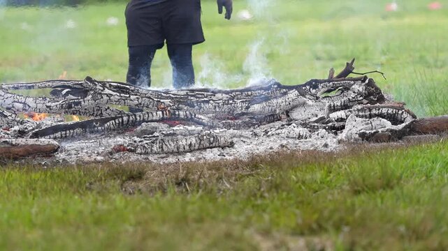 A pile of burning logs emits smoke and flames on a grassy field with a person standing nearby.
