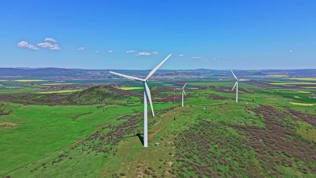 Tall wind turbines spin slowly on green hills. Fields stretch out in the background under a blue sky with few clouds. Mountains can be seen beyond the fields, showing a natural scene.