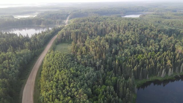Drone view looking down at a gravel road winding through a northern park forest with many small lakes. The distant trees are are hidden in forest fire smoke.
