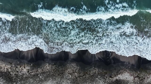 Waves flow onto the shore during high tide, reaching the sandy beach. The ocean reflects sunlight while the coastline is visible from above, showing patterns in the sand and water.