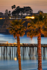 Obraz premium Seaside pier at Capitola Beach illuminated at sunset with palm tree wrapped in lights, reflections on calm Pacific Ocean water and colorful twilight sky along California coast