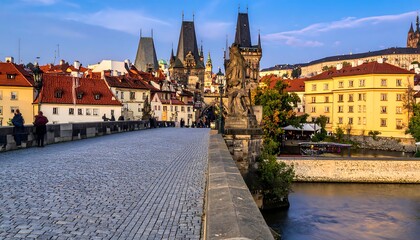 Fototapeta premium A cobblestone bridge lined with statues leads to a historic cityscape under a blue sky with scattered clouds