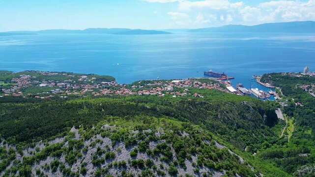 Aerial view of Rijeka, Croatia, showcasing its vibrant coastline, bustling Viktor Lenac shipyard, and lush green hills under a clear summer sky.