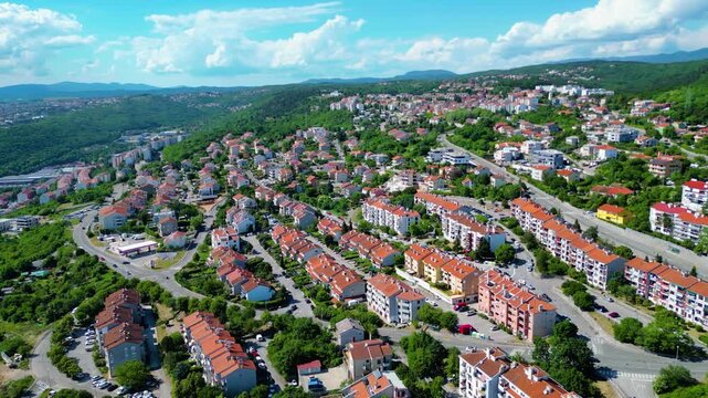 Aerial view of a vibrant residential area in Rijeka, Croatia, nestled on green hills under a bright blue sky with scattered clouds. Terracotta roofs dot the landscape. Skurinje