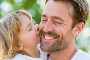 Fototapeta premium Smiling father with light brown hair receives a kiss from his young child with blonde hair, both enjoying a sunny outdoor setting with blurred greenery in the background