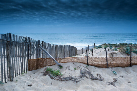 Dramatic twilight atmosphere on a deserted beach in Labenne Ocean with a traditional wooden fence and sand dunes under a dark blue overcast sky.