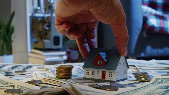 A hand gently places a tiny house amongst stacks of cash and coins, symbolizing dreams and financial goals. This scene captures the essence of investment and aspiration