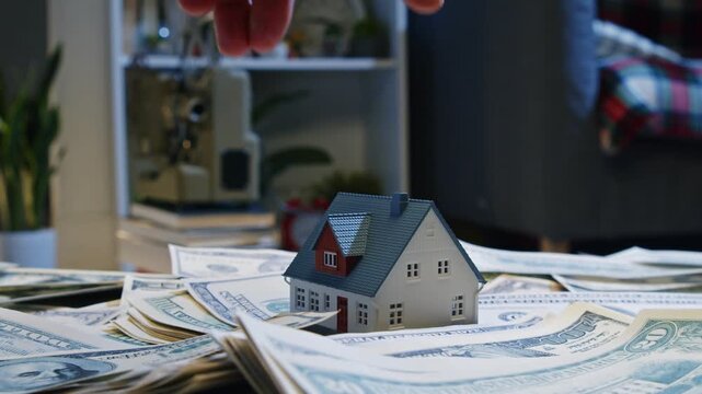 A hand gently places a tiny house on a mound of cash, symbolizing the journey of investing in real estate. Dreams of homeownership come alive in this moment